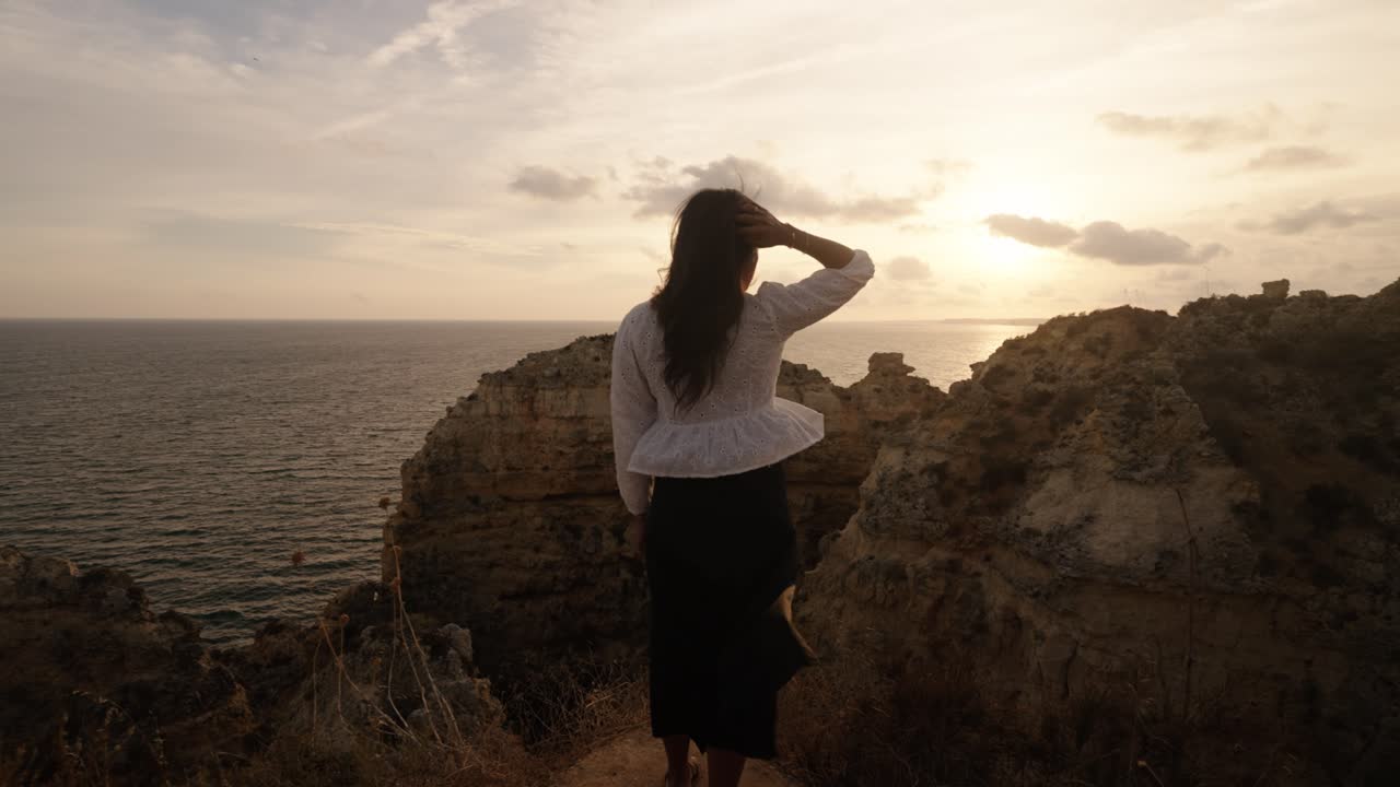 Woman stands still on Albufeira cliffs during sunset, facing the sea as golden light bathes the rugged coastline