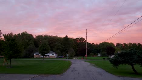 POV-shot-while-walking-over-narrow-roads-beside-green-garden-in-front-of-houses-with-red-autumn-sky-in-the-background