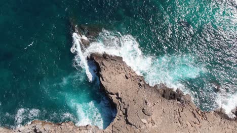 Aerial,-tilt-up,-drone-shot-of-waves-hitting-a-rocky-coast,-blue-sea,-on-a-sunny-day,-in-Mallorca,-Spain