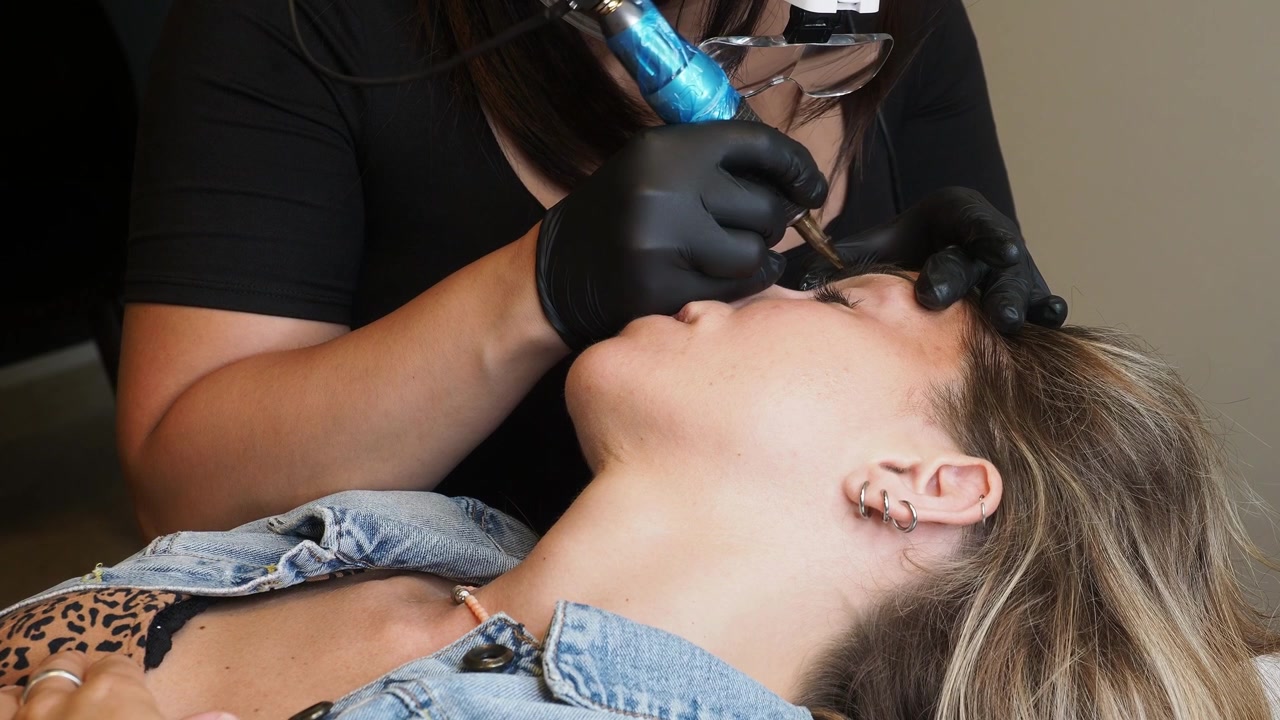 Close-up eyebrow tattooing procedure with technician wearing black gloves and using a cosmetic tattoo machine near eyebrow, with handheld tools and pigment equipment visible during beauty treatment.ey
