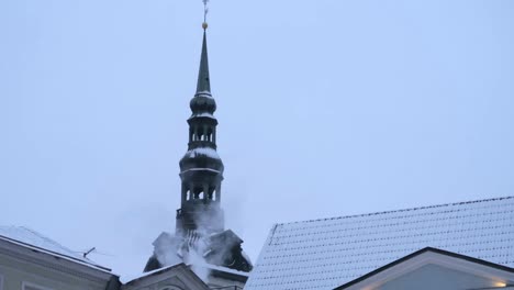 Kirchturm-Im-Stadtbild-Der-Altstadt-Von-Tallinn-Mit-Einigen-Schneebedeckten-Dächern-Und-Einem-Schornstein,-Der-Langsam-Weißen-Rauch-Bläst