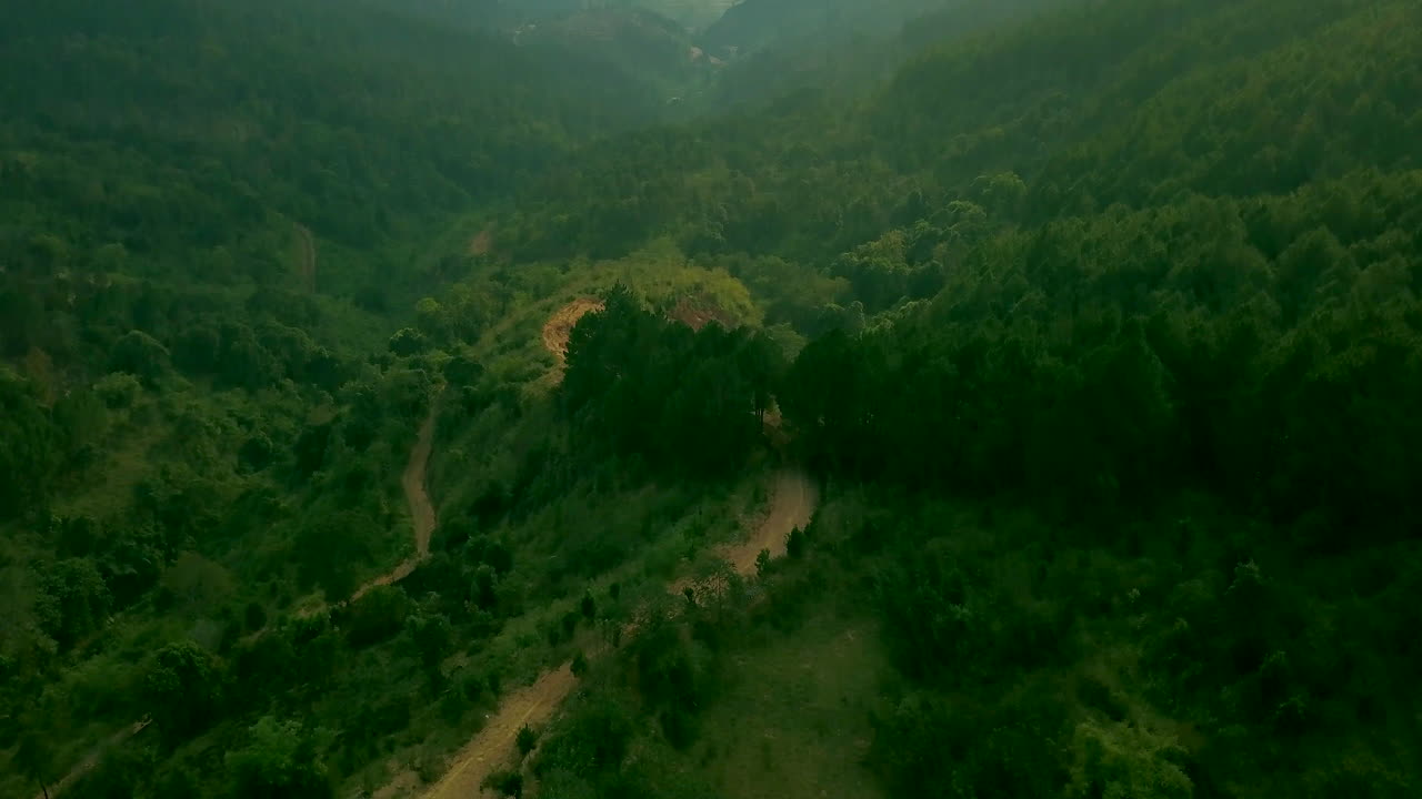 Drone shot of the road with a motorcycle and the sun shining through clouds with beautiful sun rays showing over hills of Dalat, Vietnam in Asia. Green forest and mountains and village with smoke.