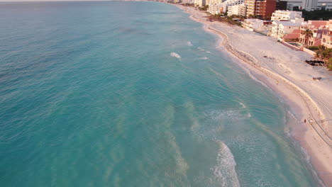 Toma-Aérea-De-Olas-En-Una-Playa-De-Arena-Blanca-Rodeada-De-Complejos-Turísticos-Y-Hoteles-En-Un-Océano-Azul-Cristalino-En-Cancún,-México