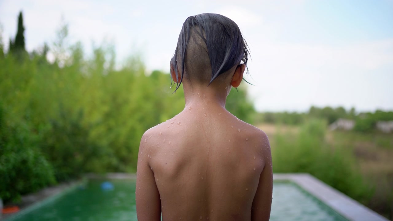 Filmed from behind, a wet boy stands looking at the swimming pool before jumping in, leaving a splash