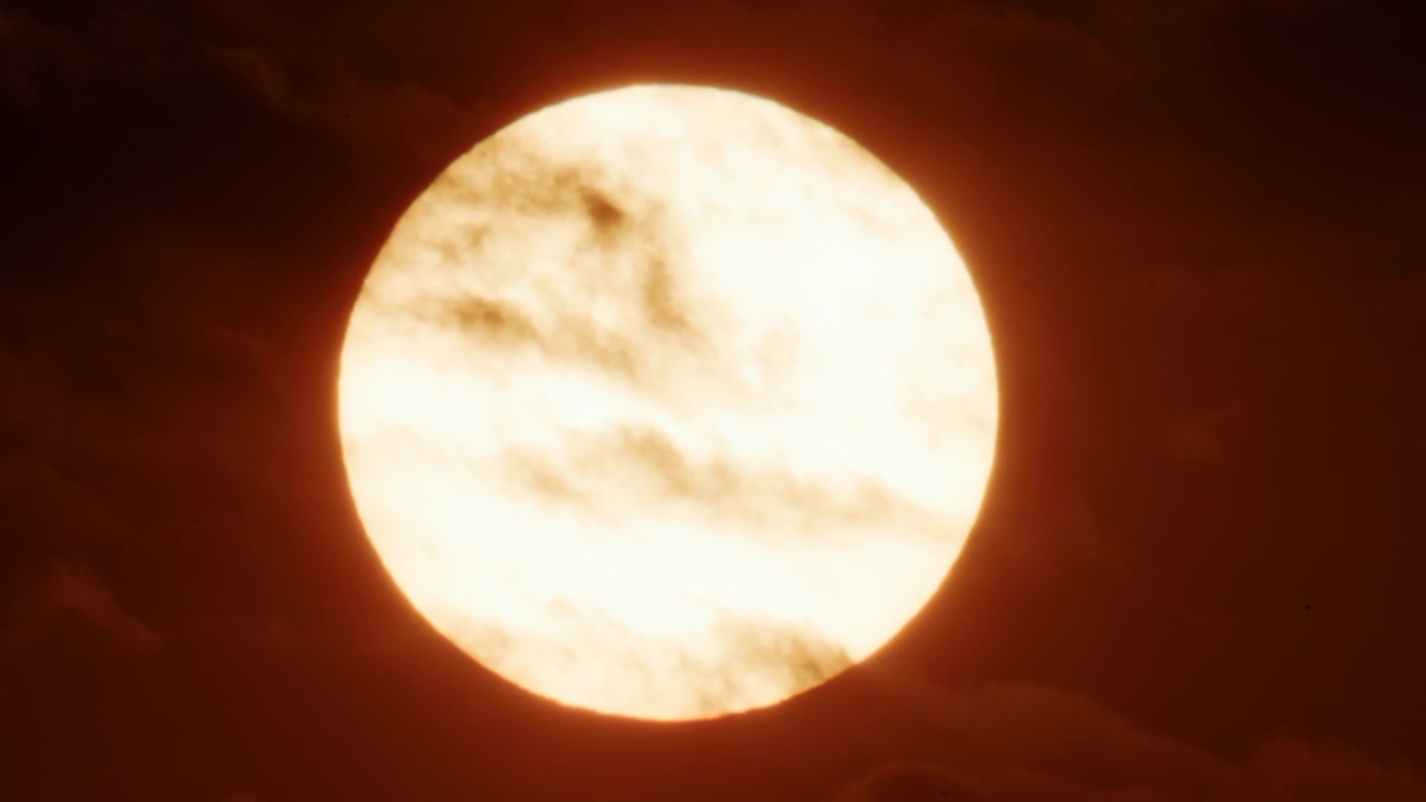Närbild av en stark sol delvis täckt av tjocka rörliga moln, som glöder i intensiva guldorange solnedgångstoner.