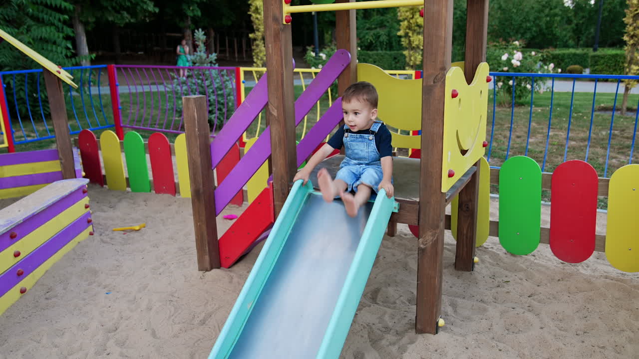 Lovely baby banging feet by the slide on the playground. Kid in a jeans romper goes down by the slide.