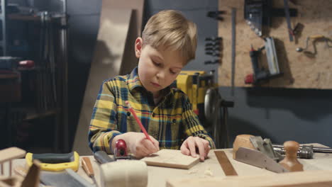 niño pequeño y lindo caucásico trabajando en un taller de carpintería y dibujando con lápiz
