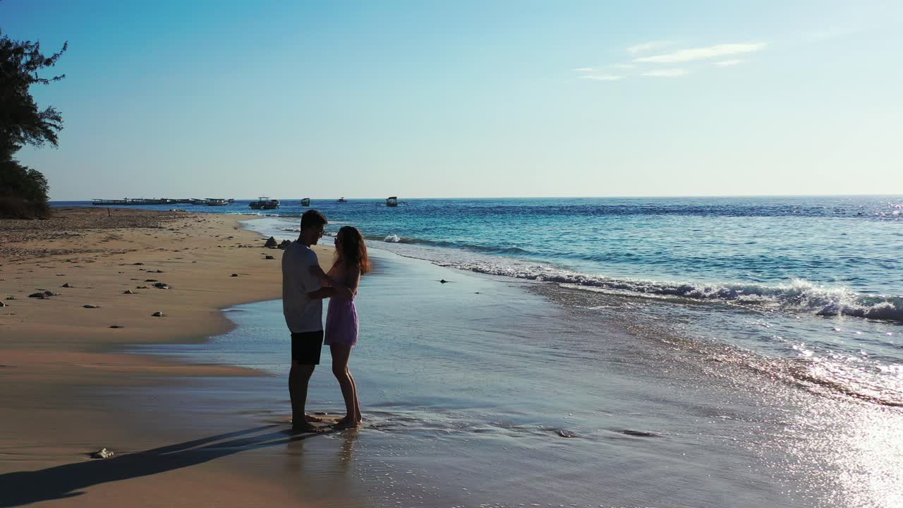 Simpatica coppia che si abbraccia sulla sabbia bagnata di una spiaggia esotica lambita dalle onde marine della laguna, con barche che galleggiano in Thailandia