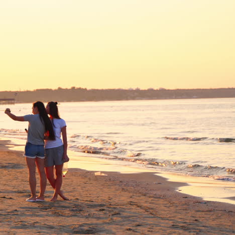 young girls are taking selfie on beach with sea at sunset. happy two friends take a photo with mobile phone in the evening on the sea shore.