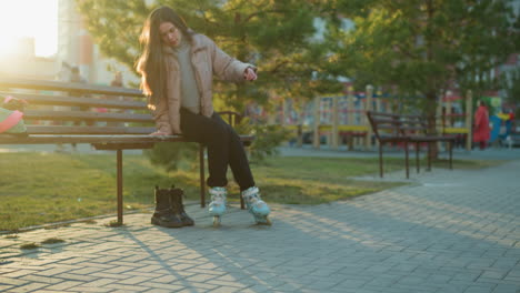 a girl in a peach jacket and black trousers is rollerblading in a park towards a bench.she is about to sit down to take off her rollerblades, with a pair of boots placed beside the bench