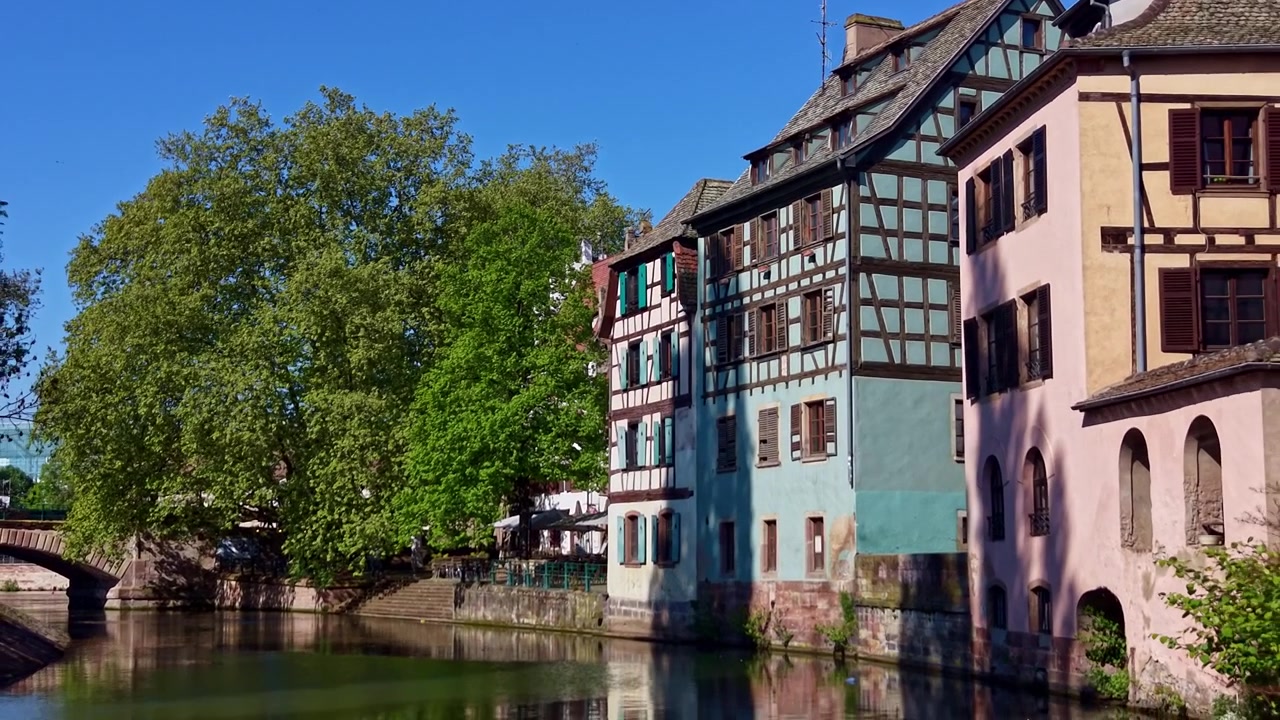 Volo di drone a bassa quota all'indietro sopra il canale del fiume Ill che mostra storici edifici a graticcio lungo il fiume nel quartiere della Petite France, Strasburgo, Francia.