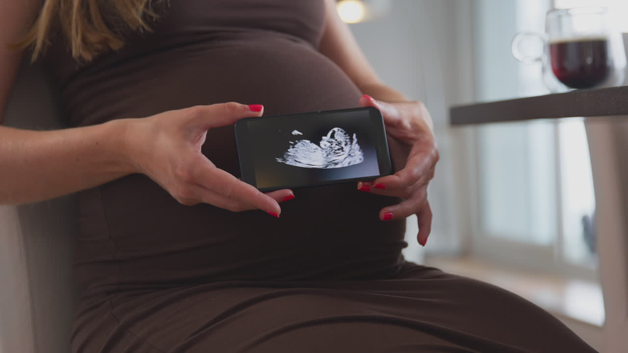Primo piano di donna incinta a casa che tiene in mano un telefono cellulare con l'ecografia del bambino