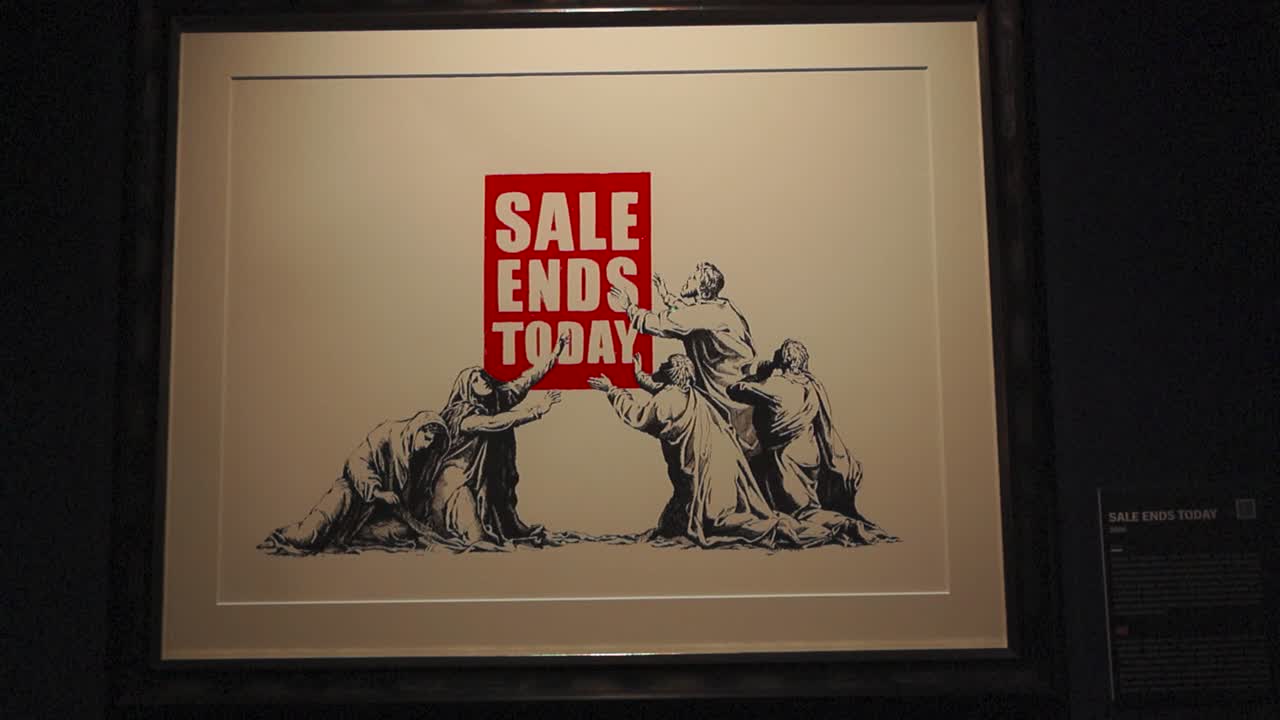 Sale ends today painting or print made by Banksy on display at his exhibition in Tallinn Estonia on a wall in black frame. The slogan is red and religious people are bowing and trying to touch it.