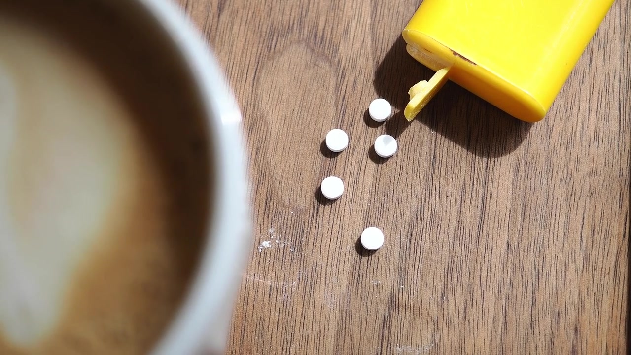 Pillole bianche rovesciate da un contenitore giallo accanto a una tazza di caffè