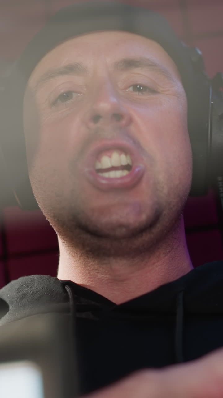 un hombre vestido de negro está usando auriculares y de pie frente a un micrófono en una habitación a prueba de sonido roja. está cantando con seriedad y moviendo su mano, transmitiendo un profundo enfoque e intensidad