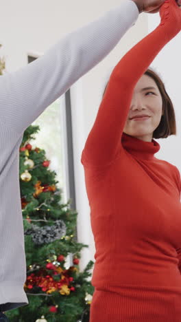 video vertical de una feliz pareja diversa con sombreros de santa bailando junto al árbol de navidad en casa