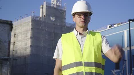 On-the-construction-site,-a-young-engineer-builder-with-glasses-puts-on-a-white-helmet-and-looks-to-the-camera