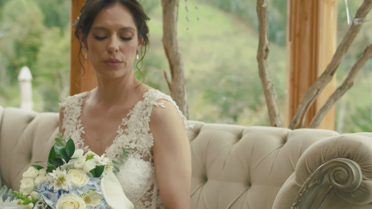 Medium shot of a bride sitting on an elegant sofa, raising her hand to look at her engagement ring. A subtle gesture full of emotion and tenderness.