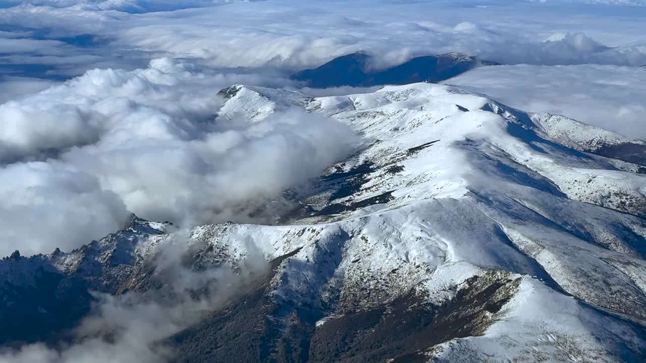 Vue aérienne surélevée de la chaîne de montagnes de Navacerrada, au nord de la ville de Madrid, prise depuis le cockpit d’un avion, partiellement recouverte de neige et de quelques nuages stratiformes de basse altitude.