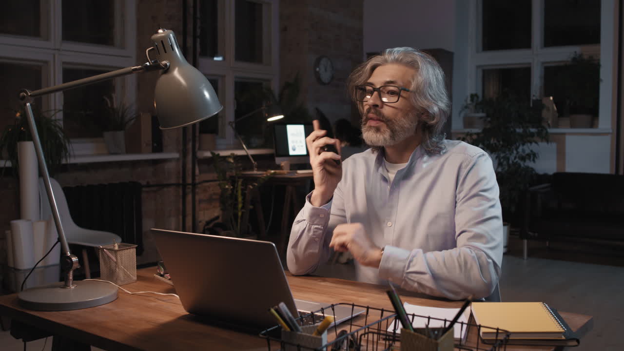 Mature Man Working Late in Home Office While on a Phone Call