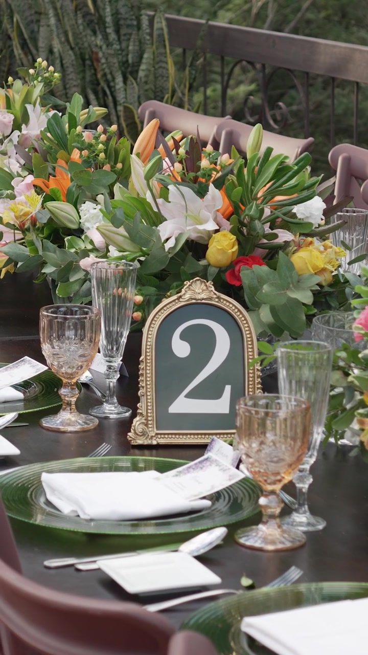 Et langt bryllupsbanketbord med et blomsterarrangement i midten og et sort skilt med tallet to. Bordet er dækket med elegant og sofistikeret service
