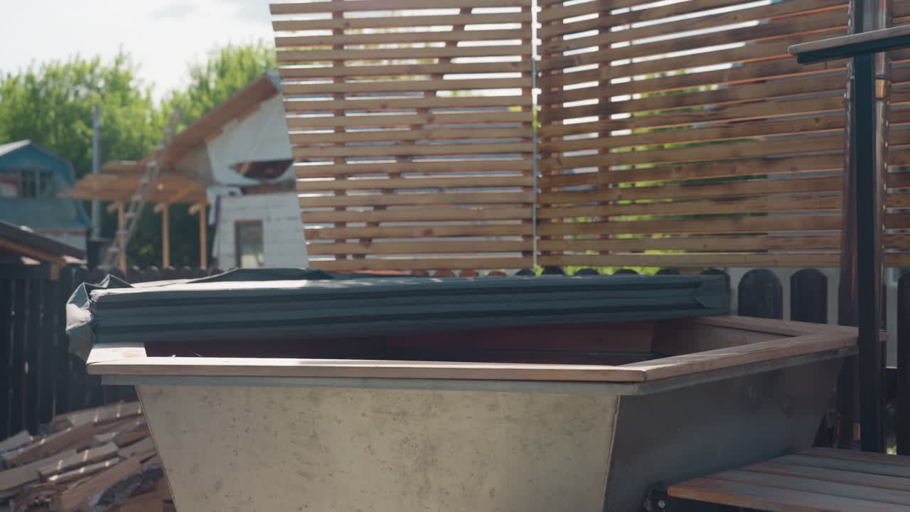 Large metal soaking tub with wooden rim and neatly folded padded cover resting along edge, surrounded by modern slatted wooden privacy fence and soft daylight casting gentle shadows in backyard