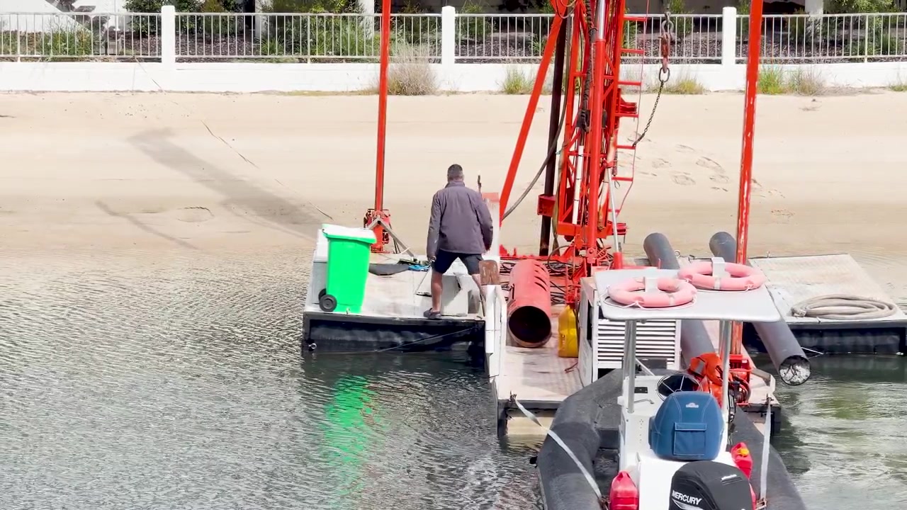 Uomo caucasico si muove lungo un pontile galleggiante di un porto turistico vicino a una struttura di gru industriale rossa.