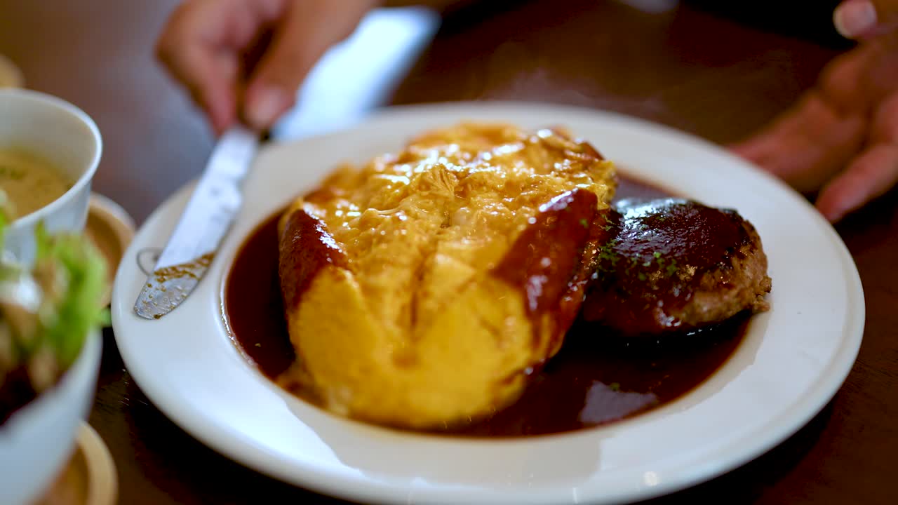 Hand slices omurice beside hamburg steak, demi-glace sauce, warm restaurant lighting, close-up view