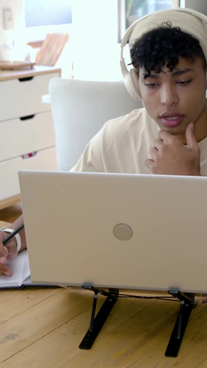 Vídeo vertical: Câmera se afastando, homem de gorro e fone de ouvido estudando no laptop e escrevendo no caderno em casa.