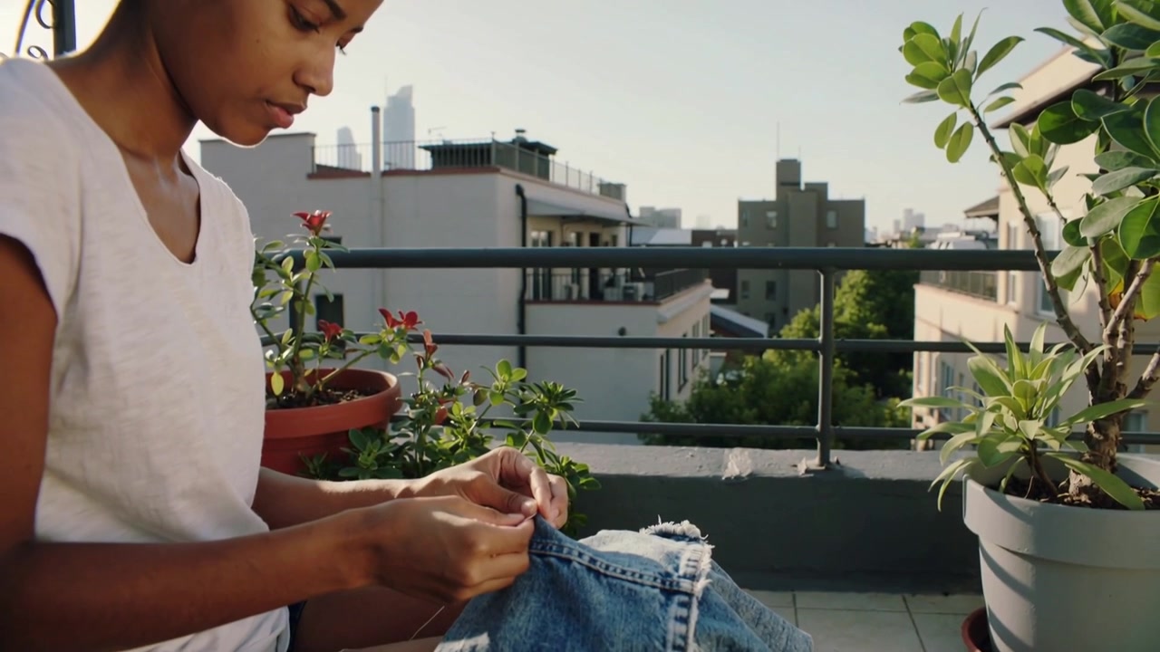 Una donna in maglietta bianca che cuce, riparando jeans sfilacciati sul balcone, usando ago e filo.