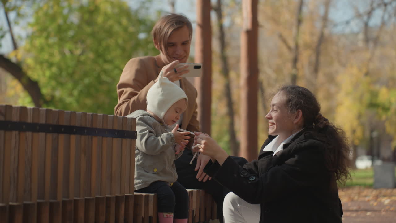 Familjestund i parken: vit förälder överlämnar småbarn, gyllene höstljus, träbänk, mormor/farmor justerar hatt, smartphone fångar äkta minne, varmt tillgivet band, mysig jacka, milda skratt.