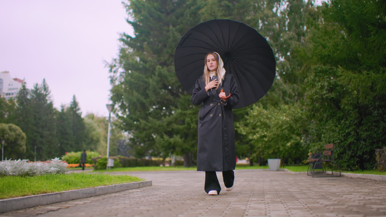 joven paseando por un parque con un paraguas mientras habla por teléfono, con un abrigo negro y capucha en un día lluvioso y nublado, rodeada de árboles verdes y bancos; escena de comunicación de estilo de vida con ambiente melancólico