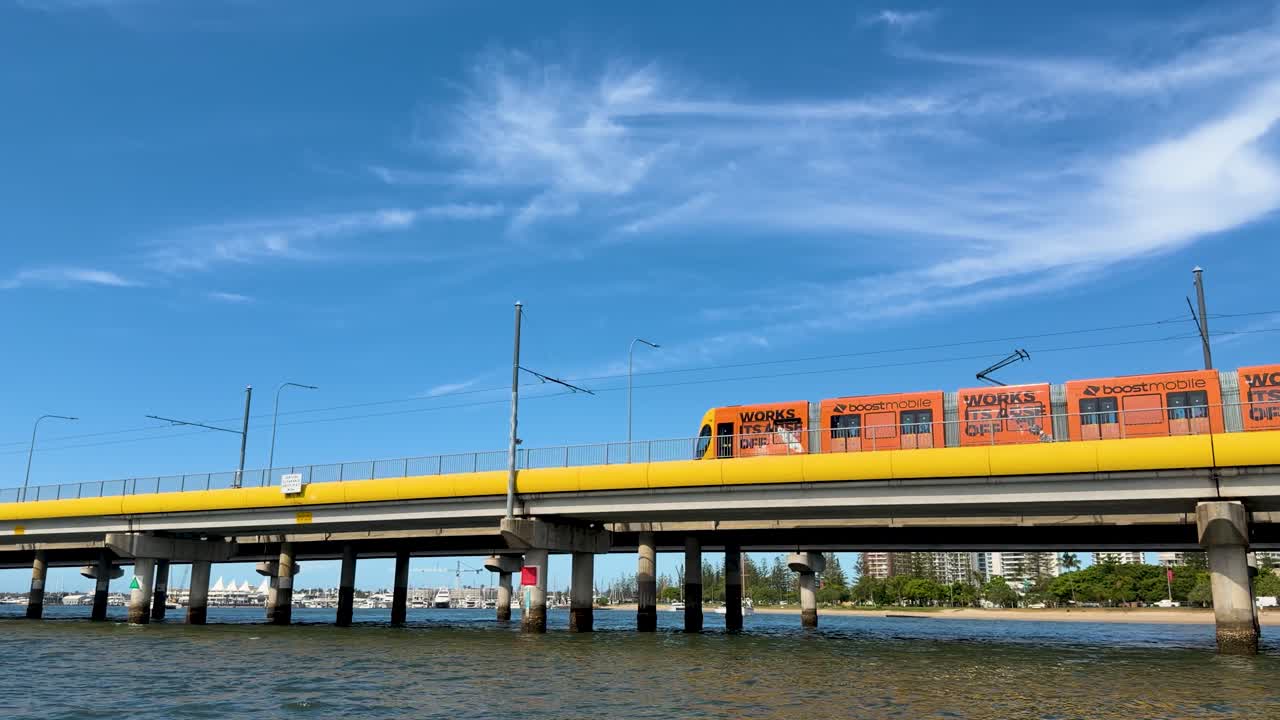 Orange light rail tram travels across yellow bridge above river, clear blue sky, wide shot
