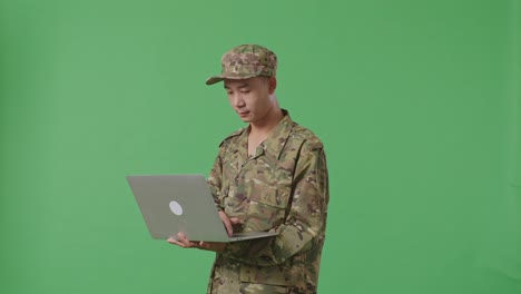side view of asian man soldier using a laptop while standing in the green screen background studio