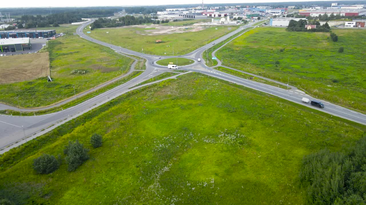 Aerial drone footage orbiting and flying closer to a large roundabout where cars, vehicles and buses drive during a summer cloudy day with houses and warehouses visible in the horizon at the back