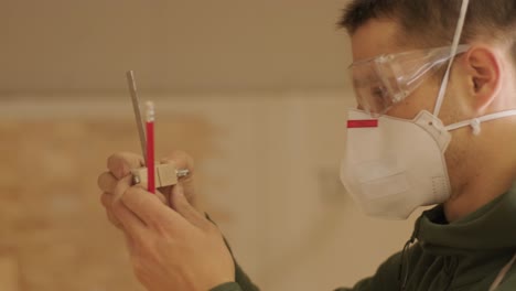 carpintero profesional trabajador con máscara y gafas sosteniendo una regla y un lápiz mientras mide una viga de madera en un taller de carpintería.