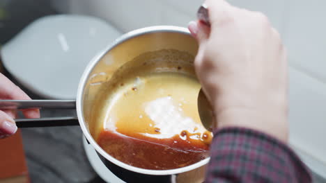 close-up rear view of person pouring rich, simmering soup from spoon into pot in kitchen, hands with polished nails carefully handle cooking process