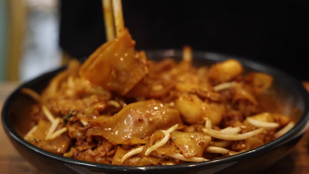 A close-up view of chopsticks lifting saucy char kway teow noodles with egg and bean sprouts from a black plate under warm, indoor lighting