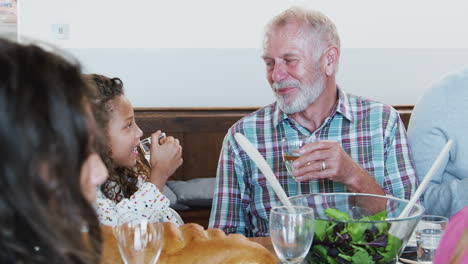 Abuelo-Y-Nieta-Hacen-Un-Brindis-Mientras-Una-Familia-Multigeneracional-Se-Reúne-Para-Comer-En-Casa