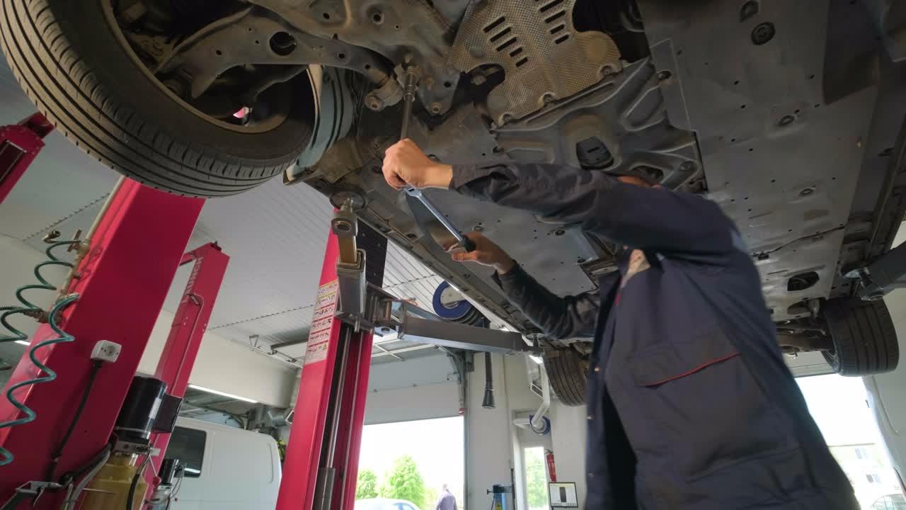 joven mecánico de automóviles en una estación de servicio de reparación inspeccionando la rueda del automóvil y el detalle de la suspensión de un automóvil levantado. vista de abajo.