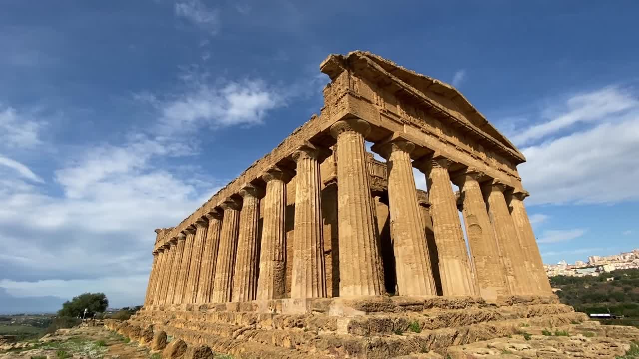 Inquadratura cinematografica da sinistra a destra delle antiche rovine greche ad Agrigento, Sicilia, che mostra le maestose colonne doriche della Valle dei Templi sotto un cielo blu drammatico