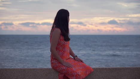 Frau-Im-Orangefarbenen-Sommerkleid-Bewundert-Den-Spektakulären-Sonnenuntergang-über-Dem-Meer-Mit-Bewölktem,-Dramatischem-Himmel