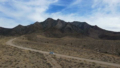 vista aérea de un auto blanco conduciendo por un camino de tierra a través del desierto en rachel, nevada con una montaña en el fondo con un cielo azul y parches de nubes blancas en un día de verano