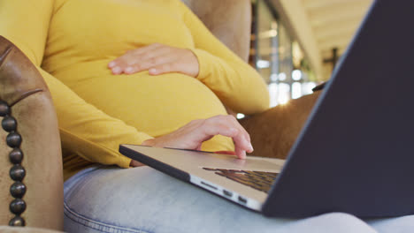 midsection of caucasian pregnant woman sitting in armchair with laptop