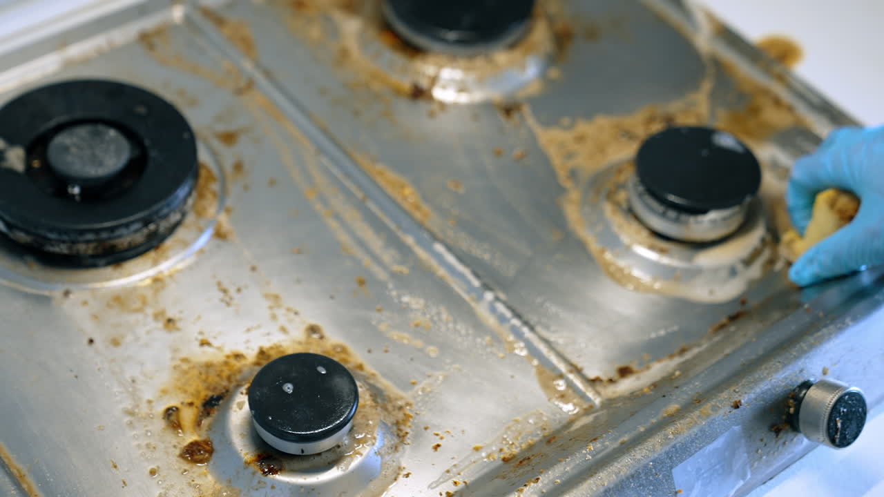 Cleaning a greasy stove in a kitchen. A person in gloves scrubs a dirty stovetop, removing grease and grime in a home kitchen on a sunny day