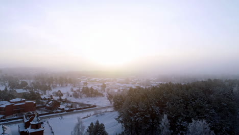 Vista-Aérea-Sobre-Las-Hermosas-Casas-Pequeñas-Al-Lado-Del-Bosque-Cubierto-De-Nieve-En-Una-Brumosa-Mañana-De-Invierno