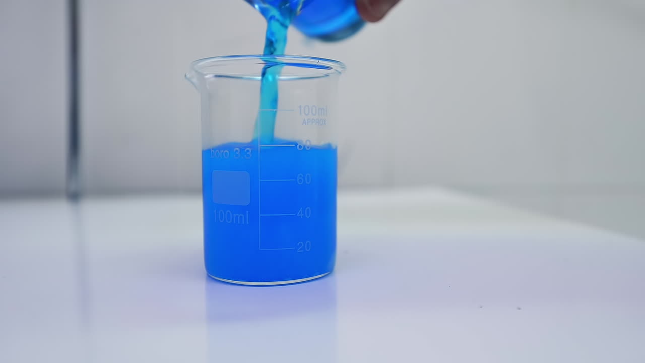 Hand of researcher pours blue liquid into the beaker with diaper filler. Blue liquid mixes up with filler and interacts with it. Close up.
