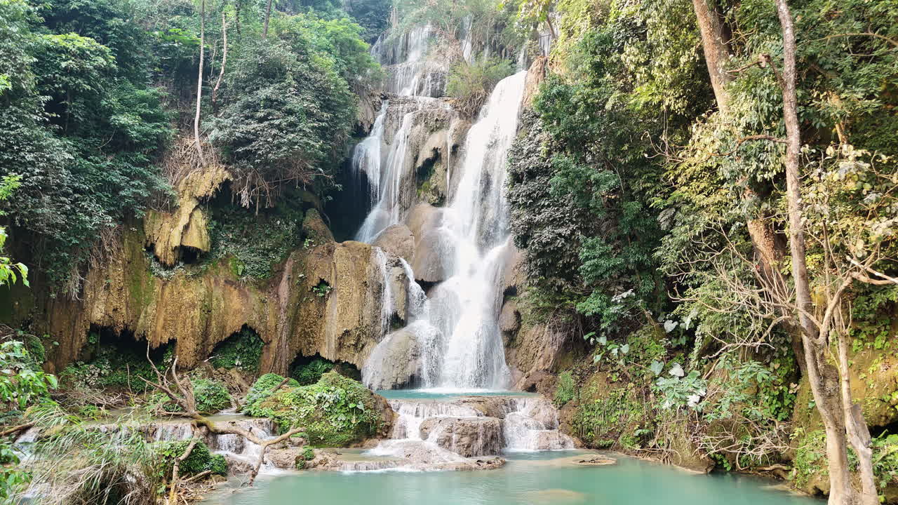 Vista aerea con drone della cascata principale di Kuang Si vicino a Luang Prabang, dove molteplici corsi d'acqua precipitano da formazioni rocciose calcaree in pozze turchesi circondate da una giungla fitta.