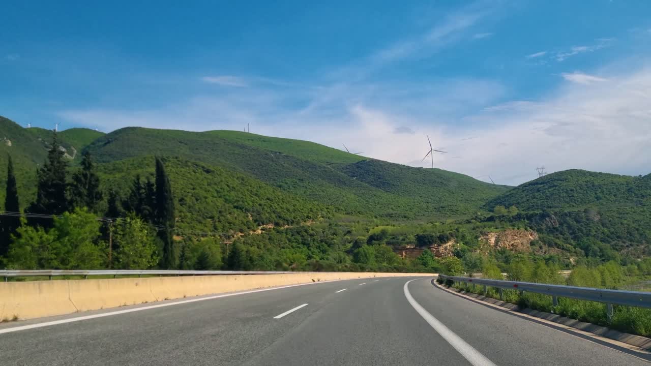 Passenger view from a car driving along a highway in northern Greece. Lush green hills dotted with solar wind turbines line the road on a sunny spring day near Serres, Macedonia