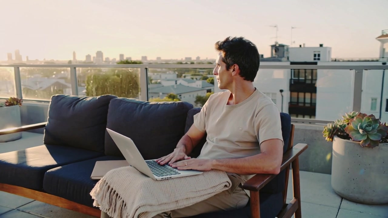 Working man in tee typing-pausing on rooftop sofa, sun drawing view gaze, laptop blanket